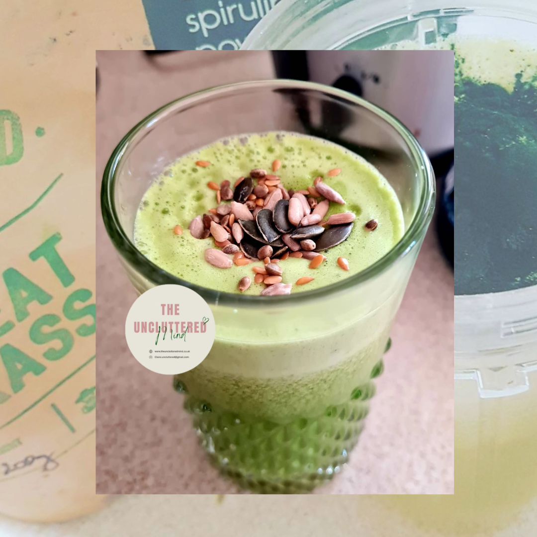 A green juice topped with mixed seeds in a textured glass, photographed in natural light. The background shows wheatgrass and spirulina powder, with The Uncluttered Mind logo overlayed in soft neutral tones.